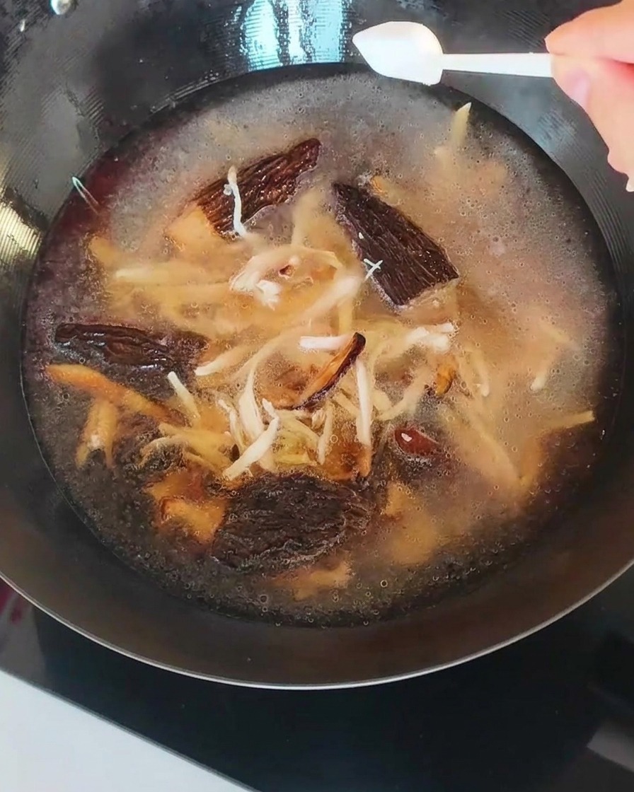 The broth in the pan, containing mushrooms and shredded chicken, being seasoned with a spoonful of salt.