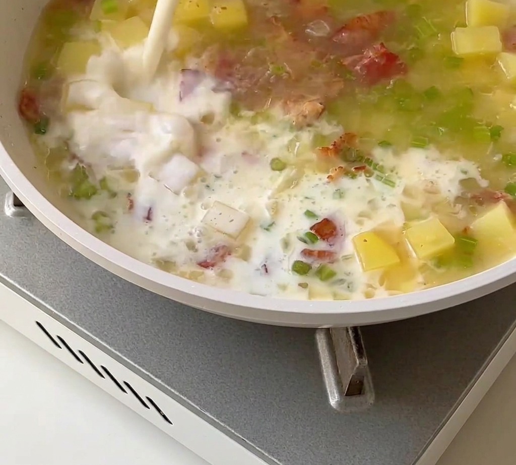 A creamy clam chowder base being stirred with milk, heavy cream, and clam meat in a pan.