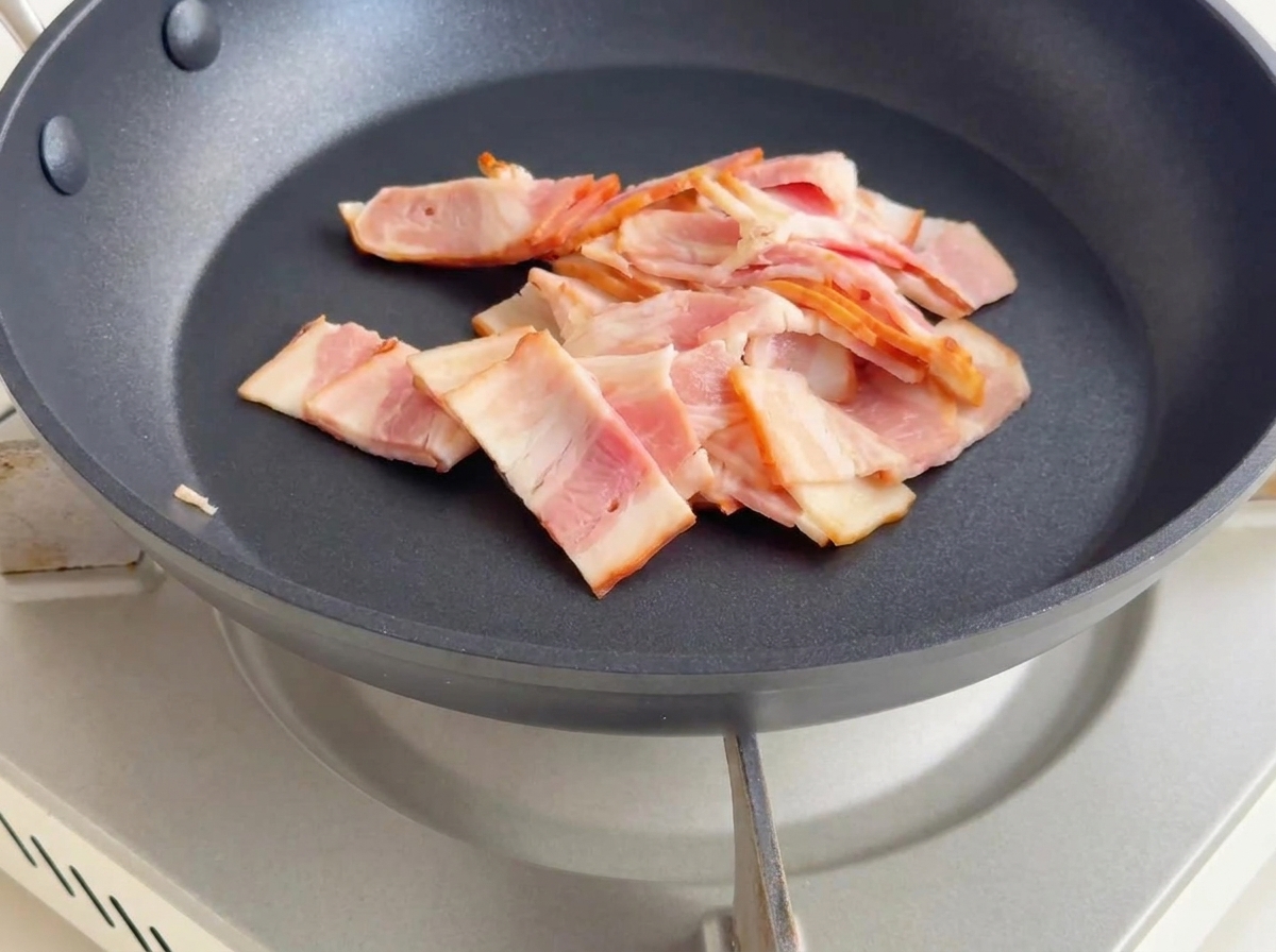 Small pieces of bacon frying in a pan, turning golden brown and releasing rendered fat.