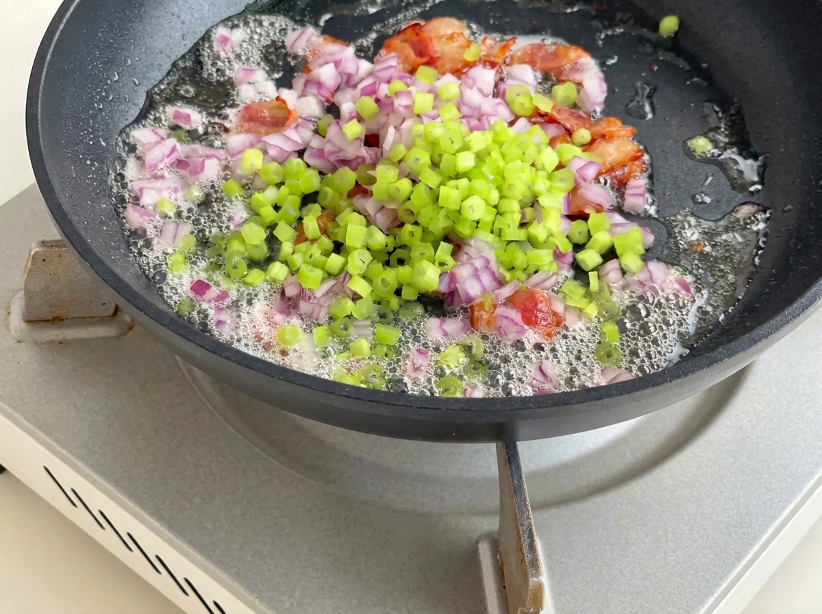 Diced potatoes, celery, and red onion being stirred into the pan with bacon and rendered fat.