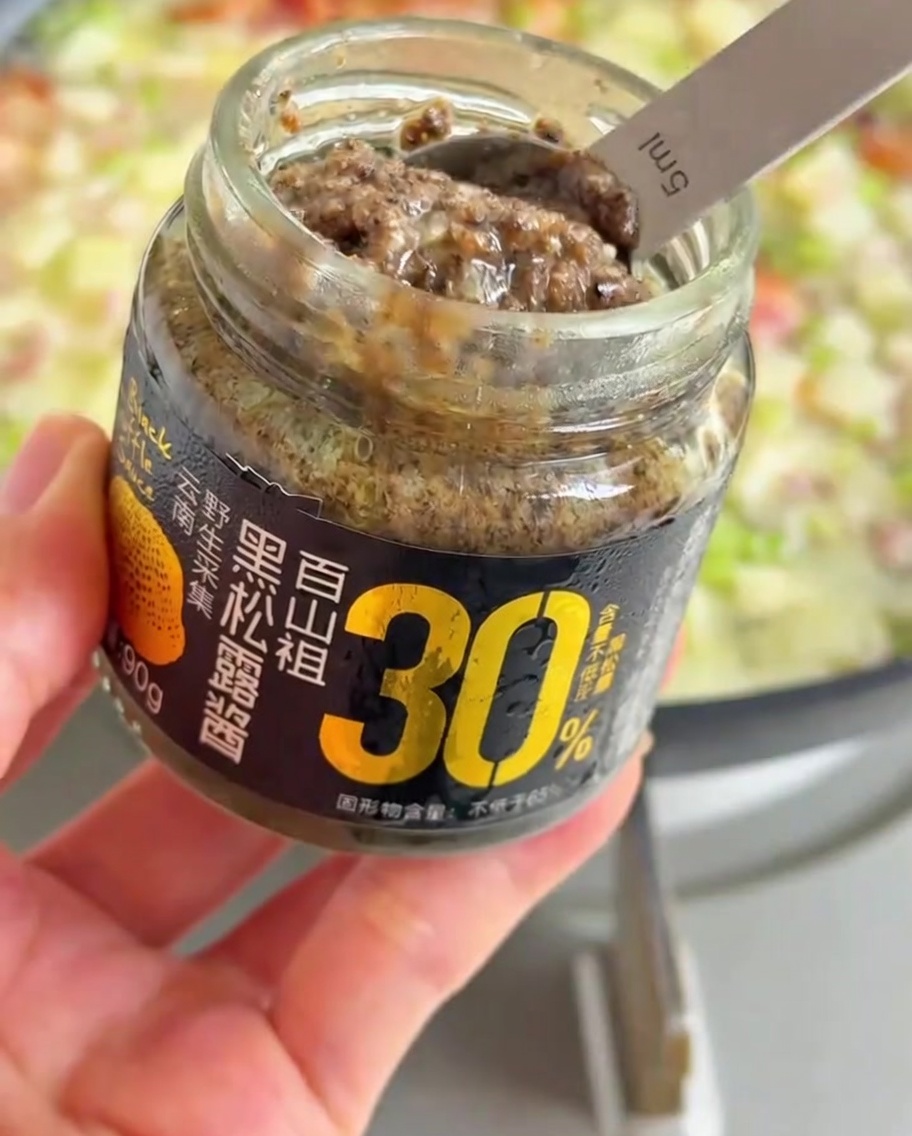 A spoonful of dark black truffle paste being added to the bubbling pan of vegetables and broth.