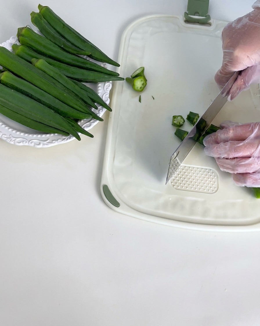 Fresh green okra pods being trimmed on a white cutting board.