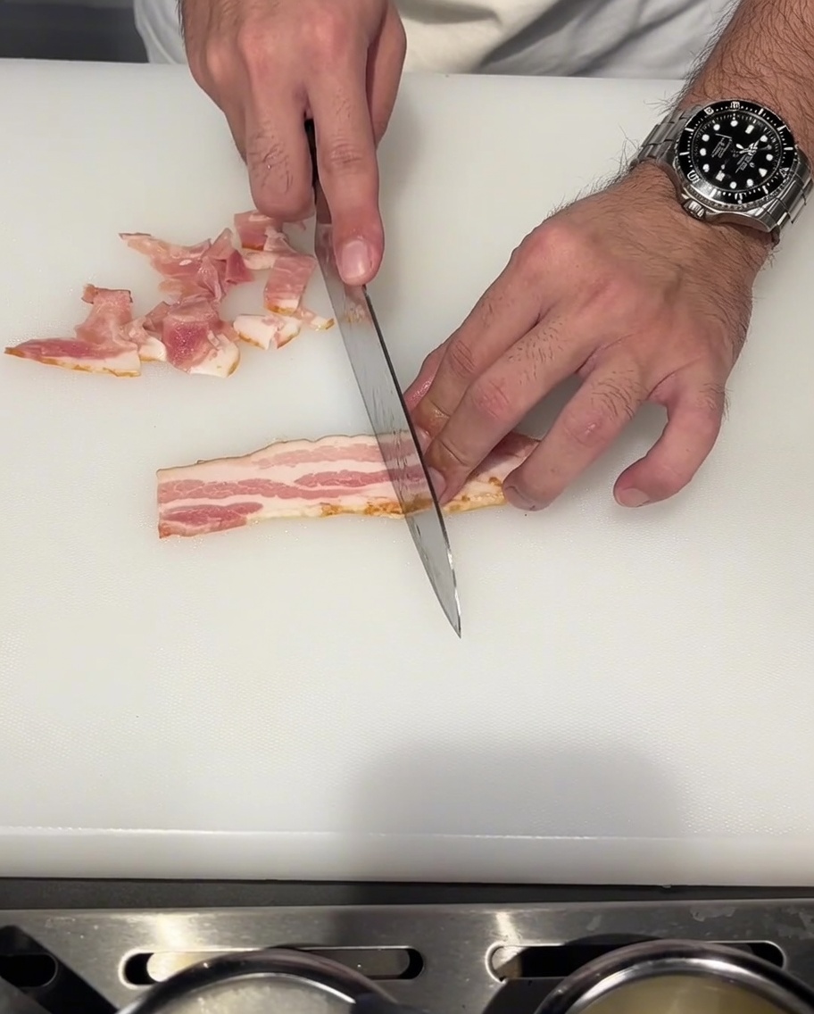 Bacon strips being sliced into small pieces on a white cutting board.