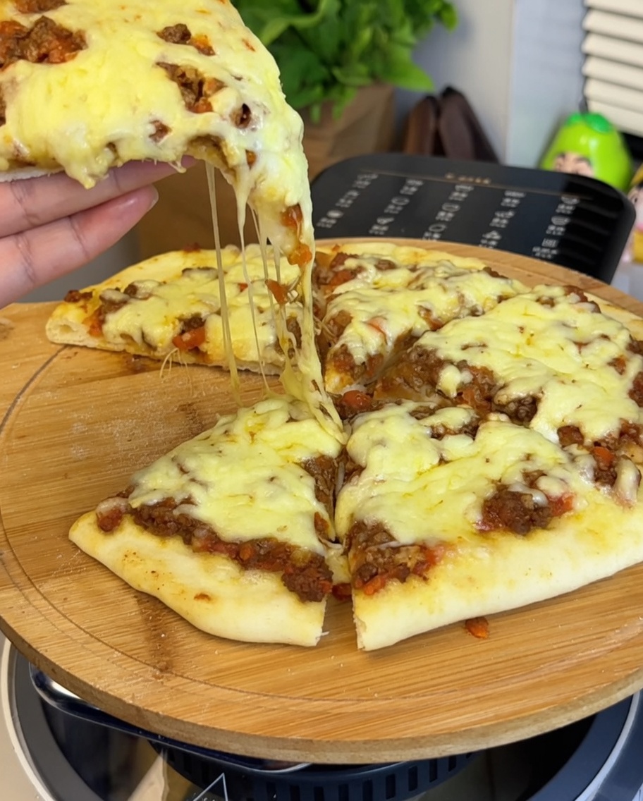 A close-up of the tomato beef sauce being spread over the pizza crust, with a hand holding a piece of pizza in the foreground showing melted cheese.