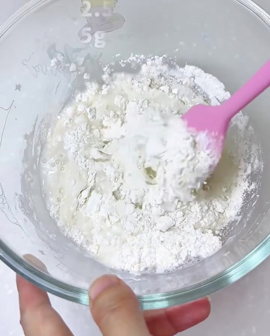 Ingredients for pizza dough being combined in a large glass bowl with a silicone spatula.