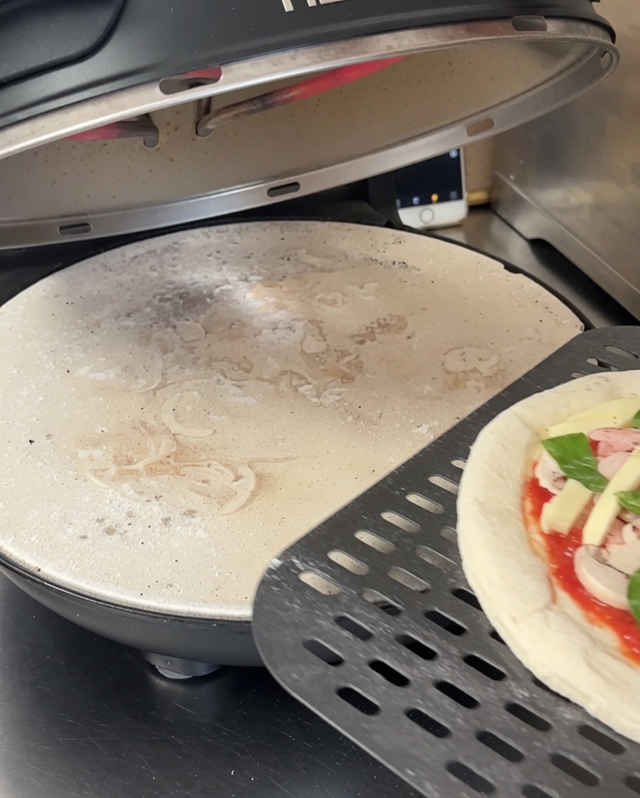 Flattened pizza dough resting on a black perforated pizza stone inside a pizza oven.