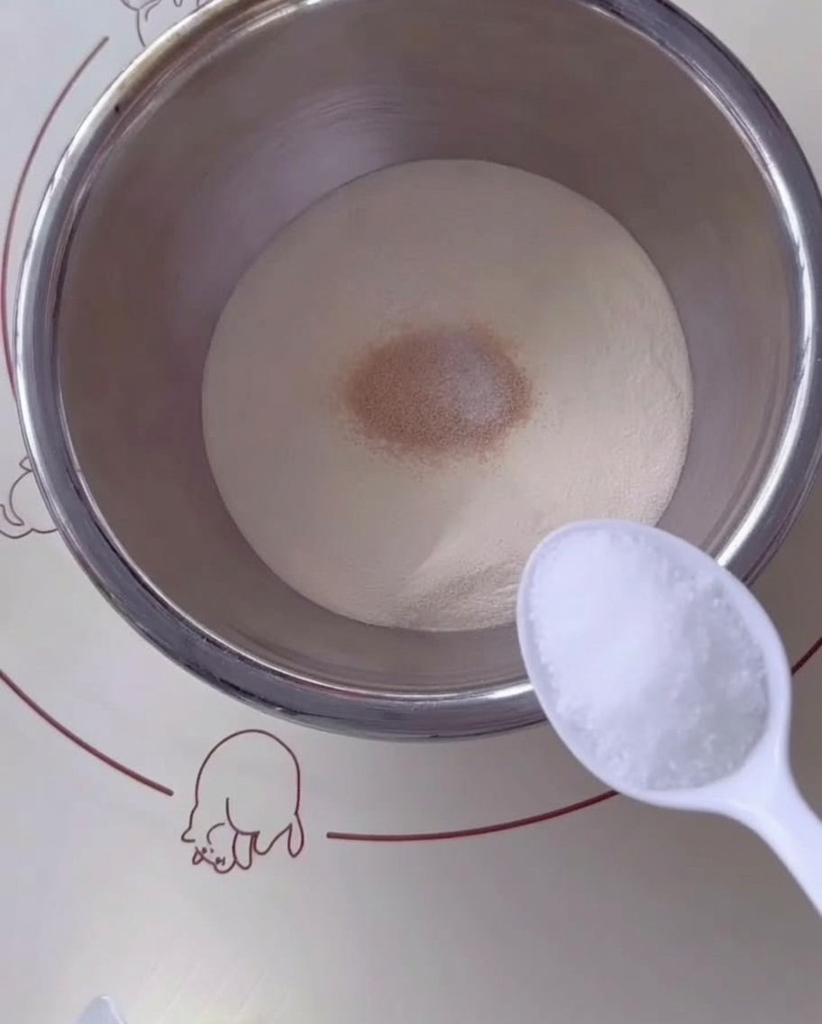 A spoonful of sugar being added to the flour and yeast mixture in a stainless steel bowl.