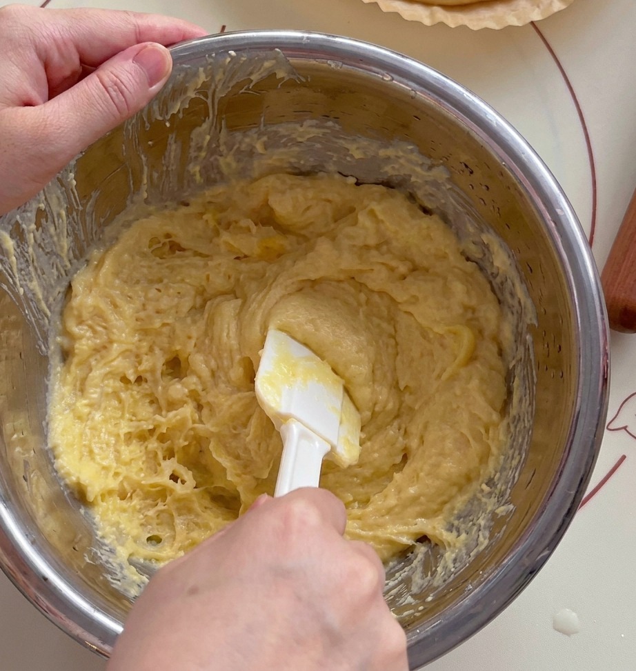 A close-up of a white spatula mashing bright yellow durian flesh in a metal mixing bowl.