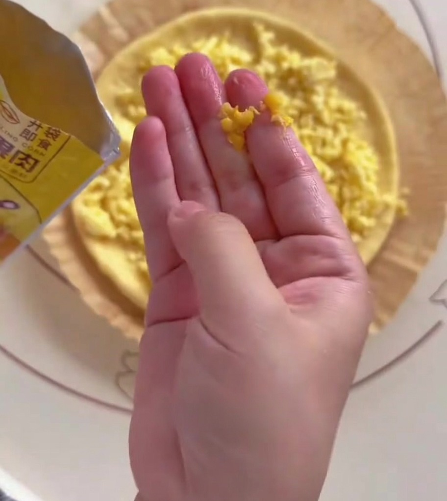 A hand scattering yellow corn kernels onto half of a round pizza dough base.