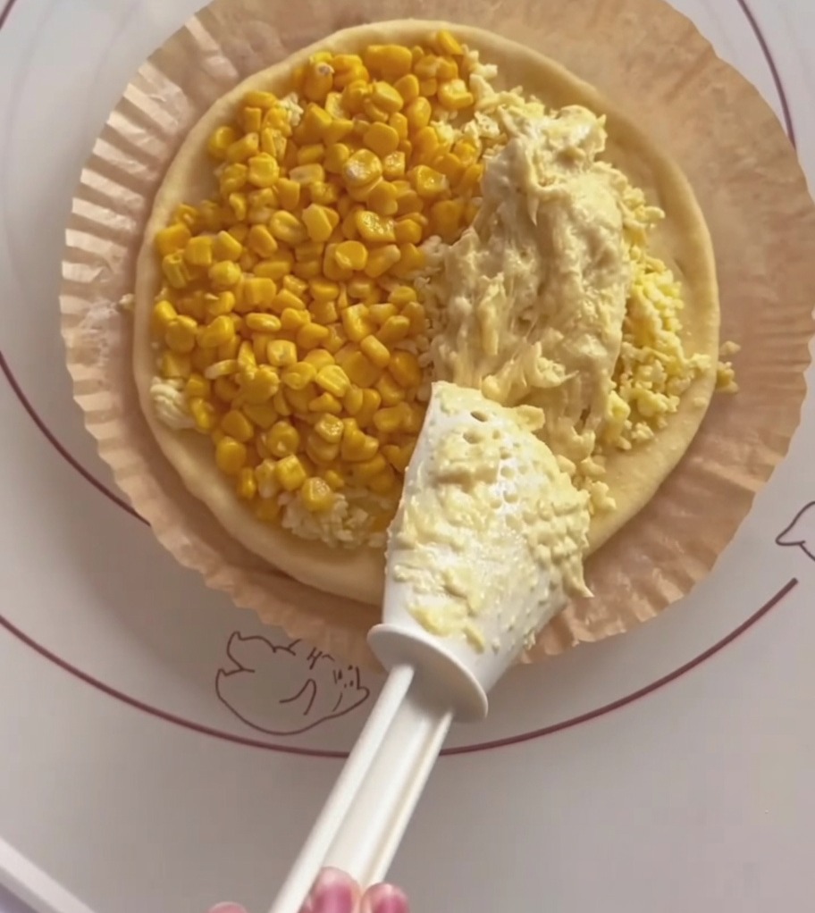 A white spatula spreading creamy yellow durian paste onto one half of a pizza base, with corn already on the other half.