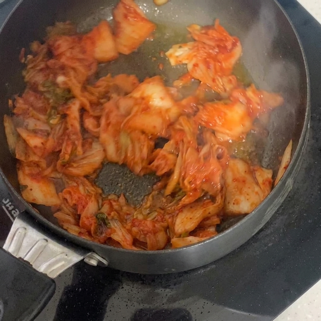 Sour kimchi pieces sizzling and being stirred in a hot frying pan with cooking oil.