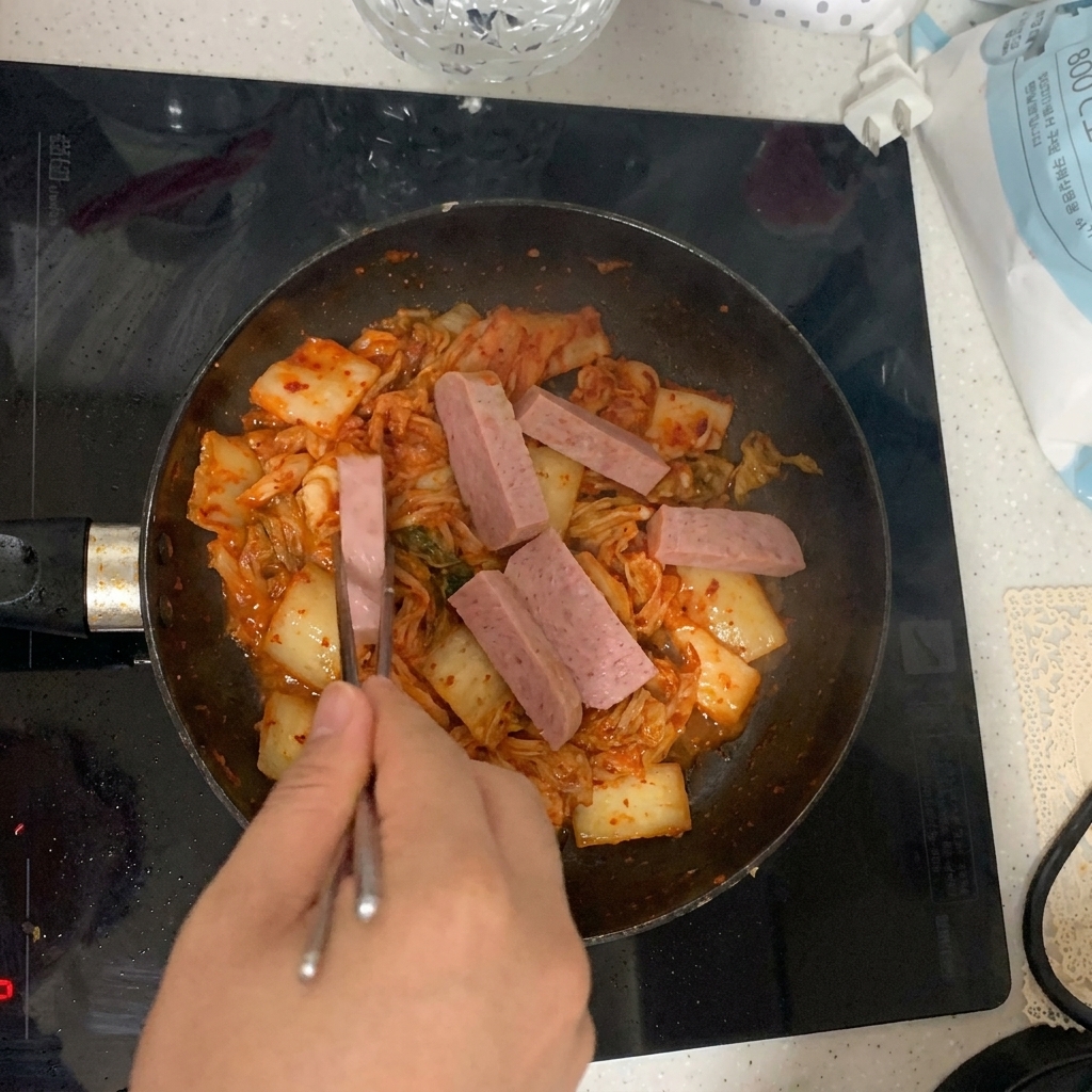 Slices of luncheon meat being placed into the pan alongside the stir-frying kimchi.
