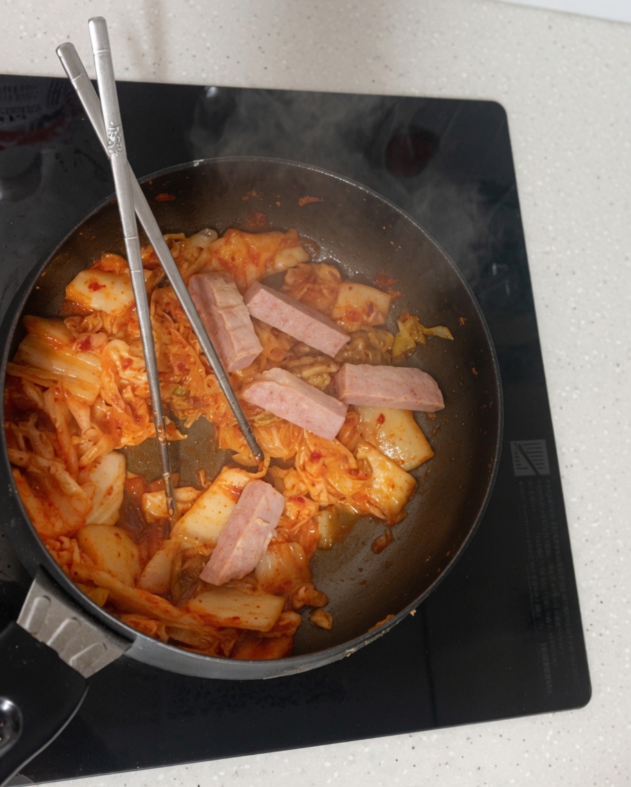 Water being poured from a bowl into the pan containing the stir-fried kimchi and luncheon meat.