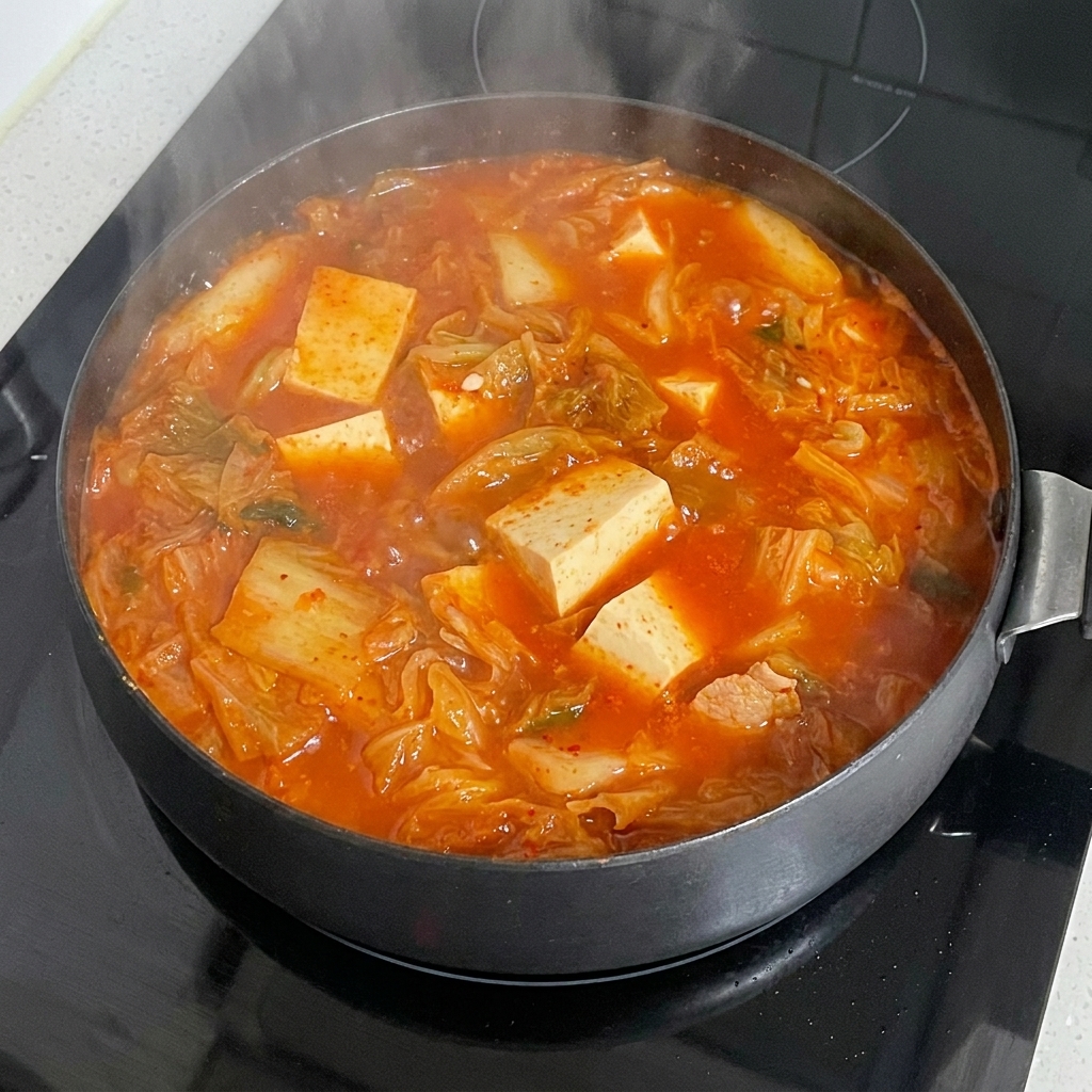A block of ramen noodles being submerged into a bubbling, vibrant red kimchi stew with tofu and other ingredients.