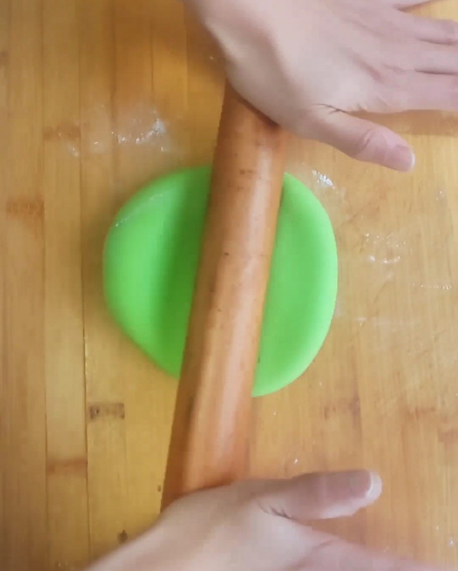 Two hands using a wooden rolling pin to flatten bright green dough into a circle on a wooden board.