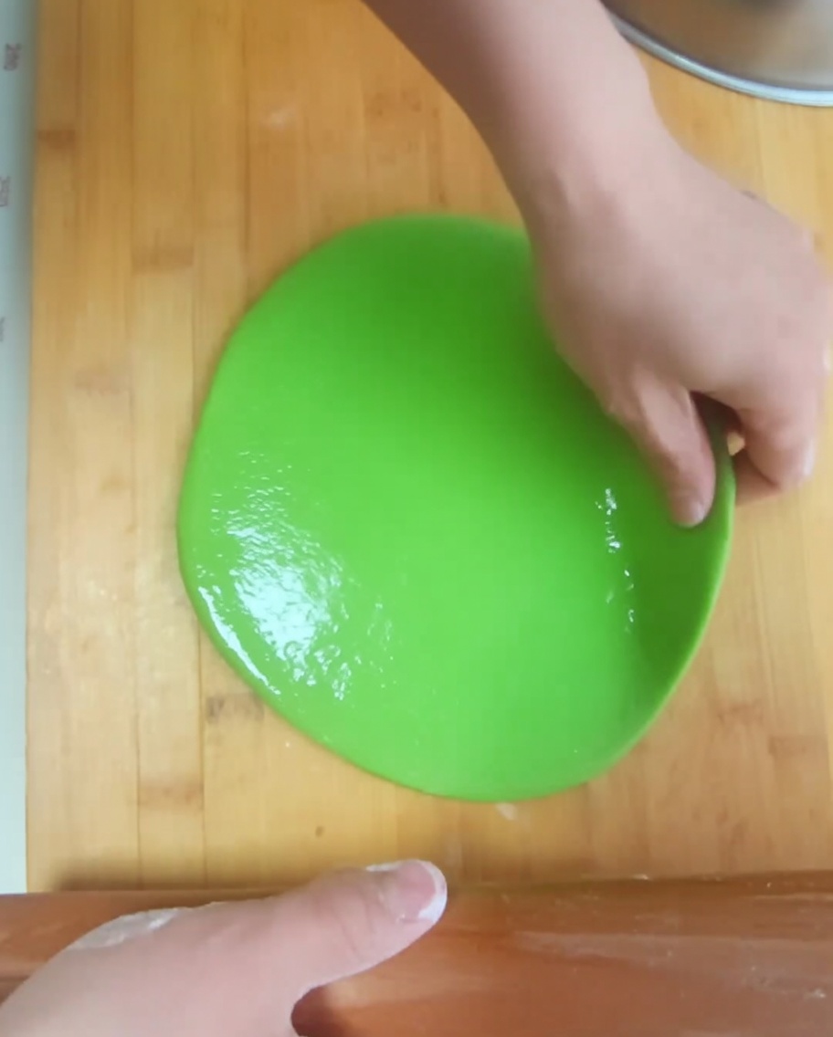 A hand spreading a shiny layer of oil over a flattened green dough circle.