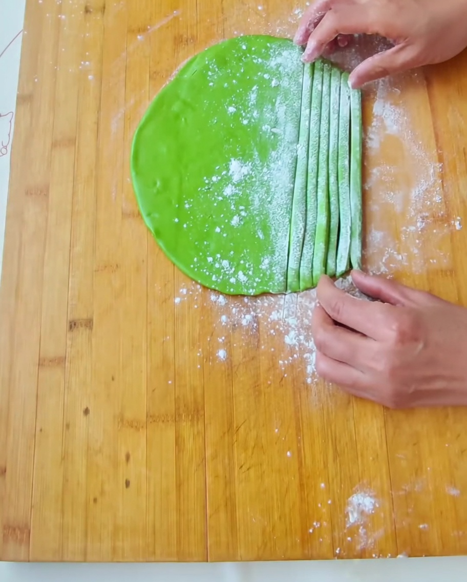 A hand guiding freshly cut strips of green dough on a flour-dusted wooden board.