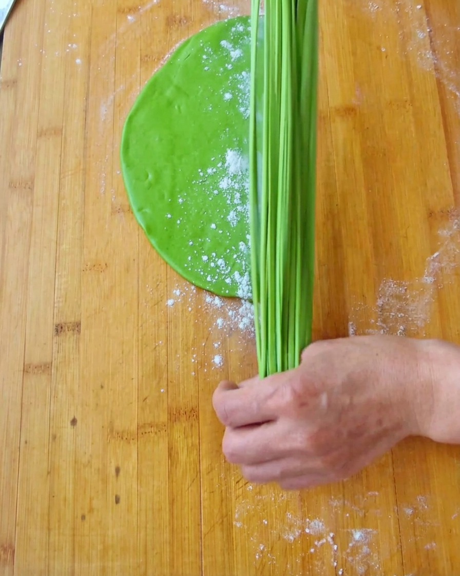 A hand holding the ends of bright green dough strips and stretching them vertically into long noodles.