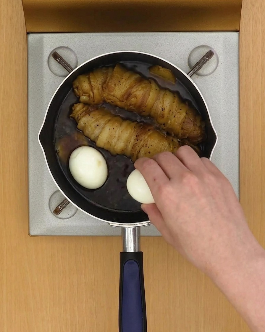 Two soft-boiled eggs being added to a pan containing pork rolls and braising liquid.