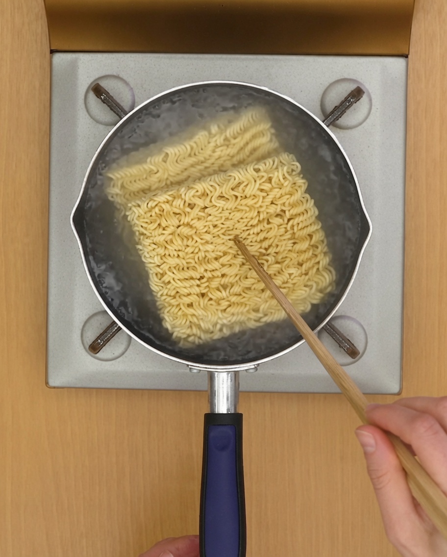 Instant noodle blocks cooking in a pot of boiling water with chopsticks visible.