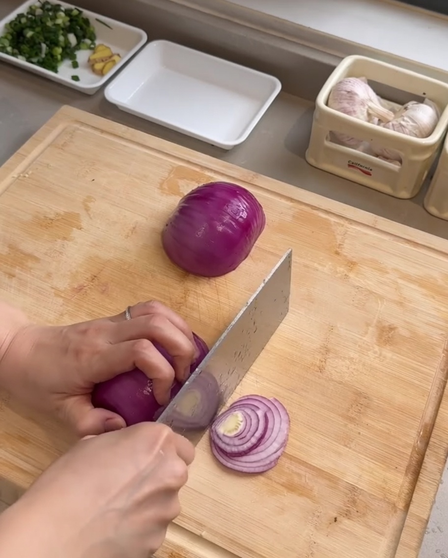 A chefs knife slicing a red onion into thin strips on a wooden cutting board.