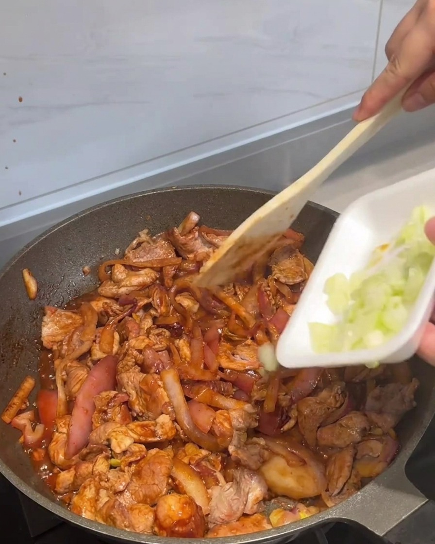 Sliced scallions being added to a pan of cooked, saucy stir-fried pork and onions.