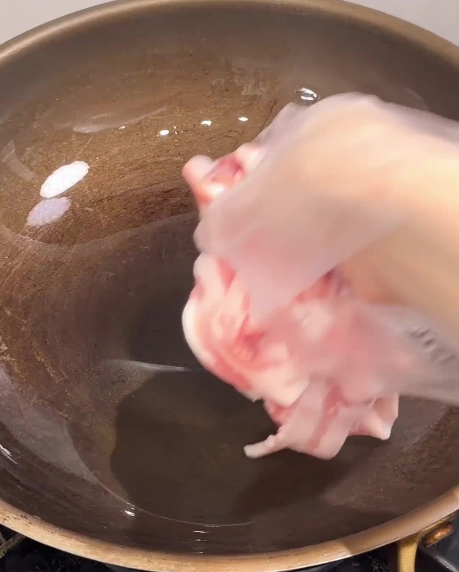Thin slices of raw pork belly being dropped into a hot, empty frying pan.