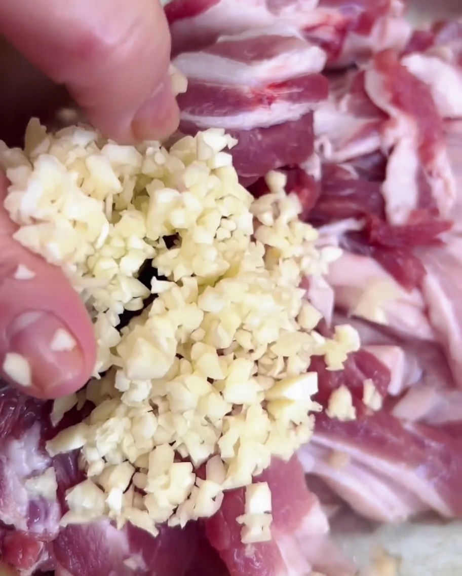 Minced garlic being poured from a hand into a metal bowl containing sliced pork.