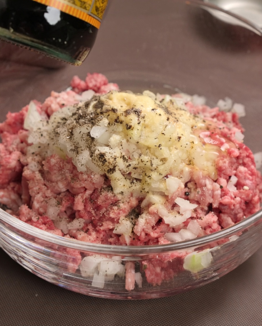 Worcestershire sauce being poured over the meat, diced onions, and seasonings in a bowl.
