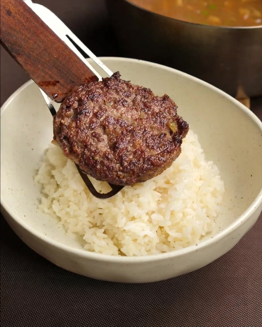 A cooked hamburger patty being placed by a wooden spatula onto a serving bowl of white rice, with a gravy pan visible in the blurred background.