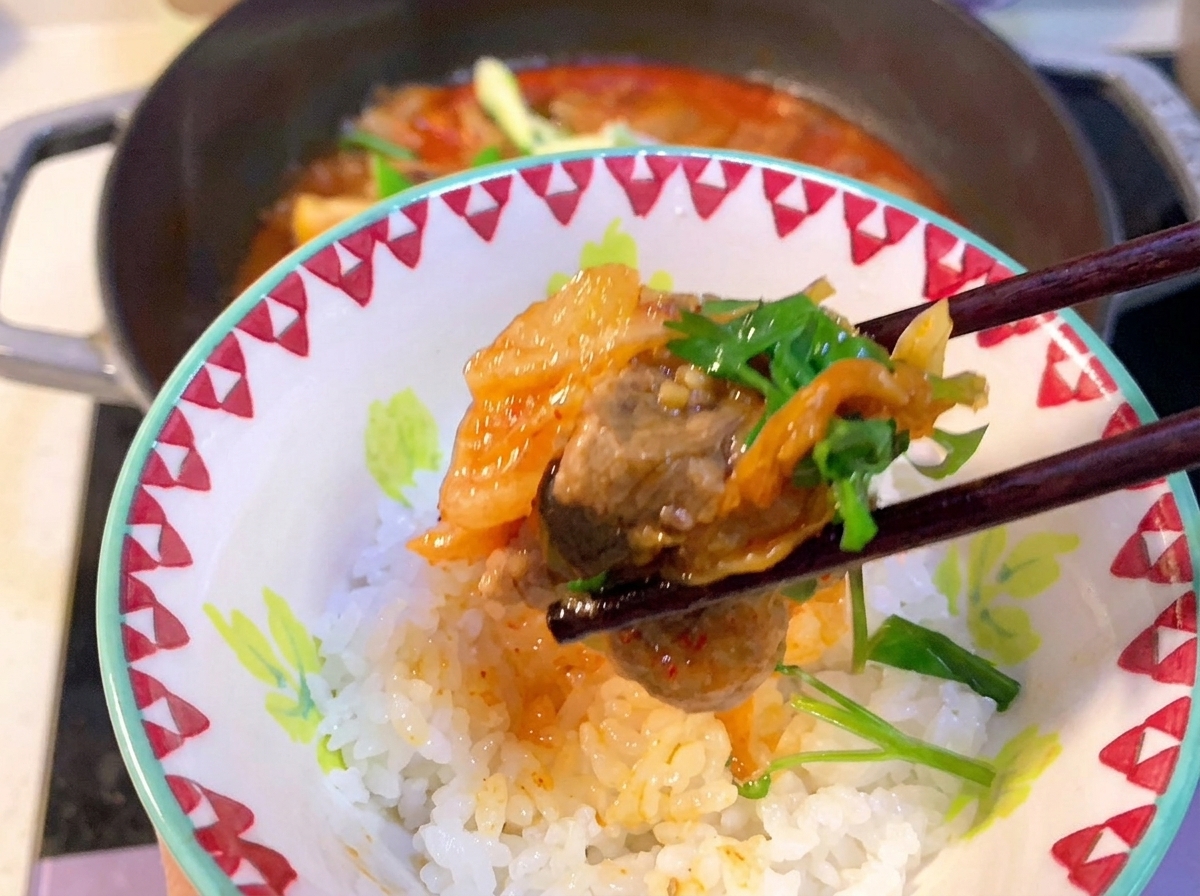 A close-up view of a spoon stirring red Korean chili paste into the bubbling kimchi and pork rib stew.
