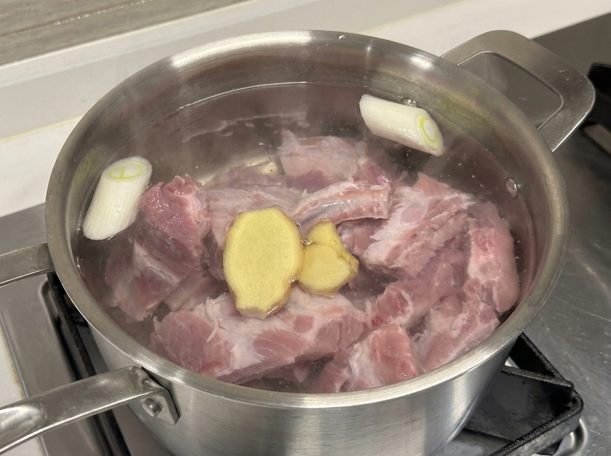 Pot containing raw pork ribs, green onion, and ginger slices with cooking wine being added.
