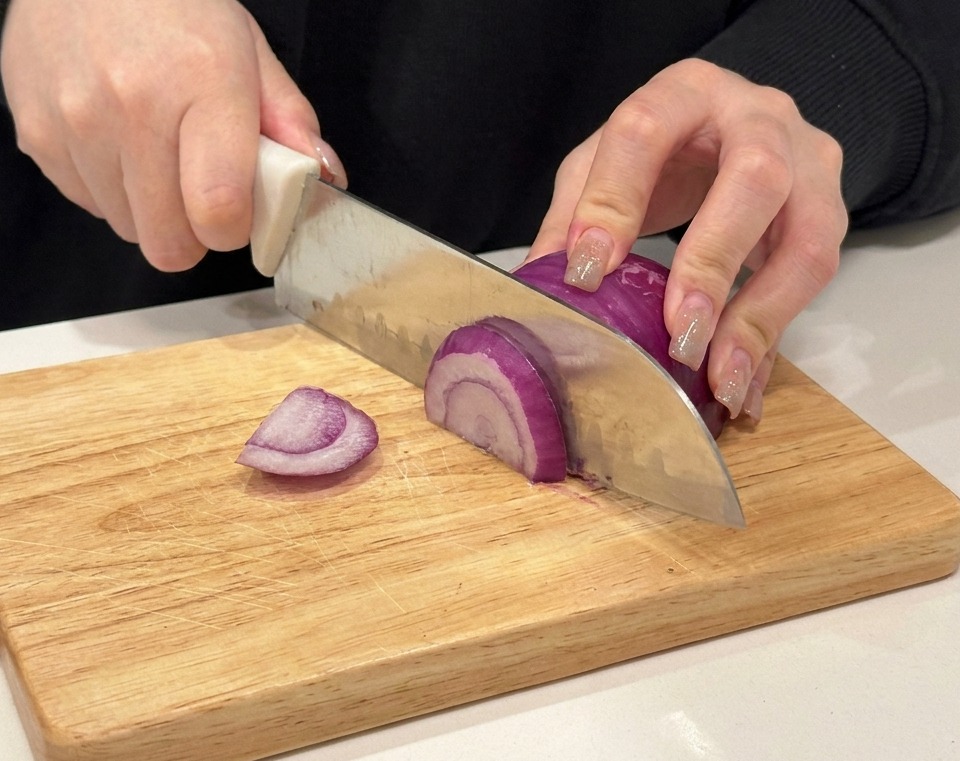 Hands slicing a red onion on a wooden cutting board.