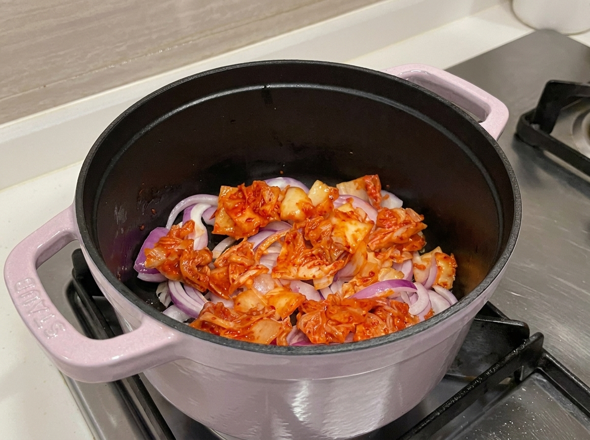 A close-up view of vibrant red kimchi and onions being sautéed in a pot, releasing their aromatic juices.