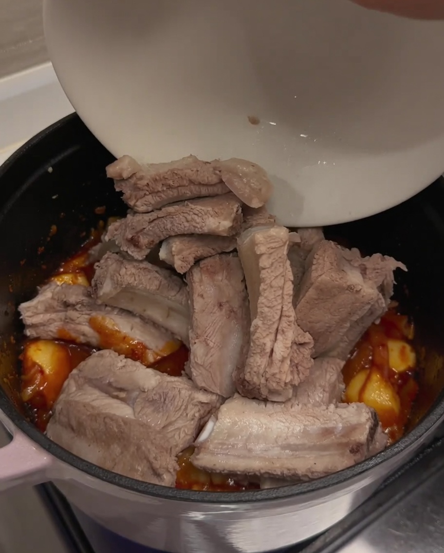 Pre-blanched pork ribs being added from a white bowl into the pot containing the seasoned, aromatic kimchi base.