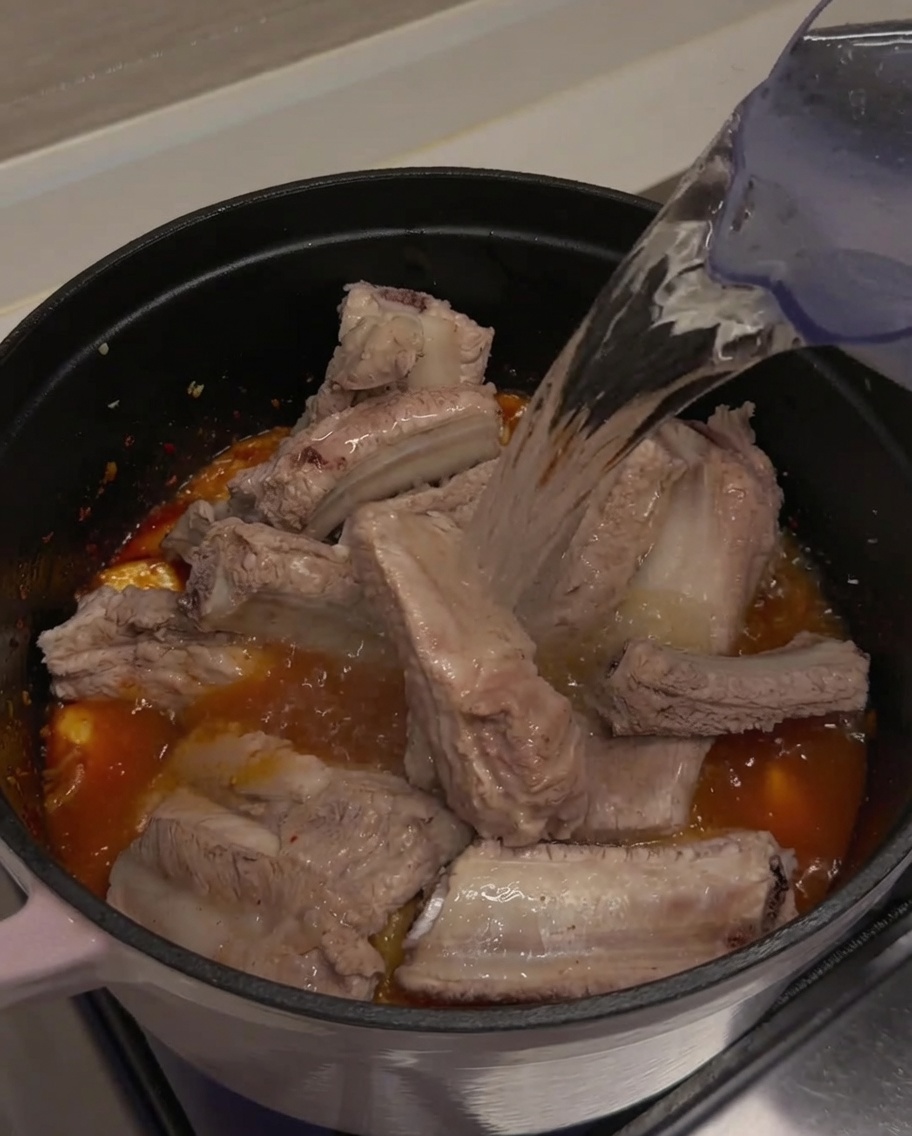 Clear water being poured from a pitcher into a pot, completely covering the pork ribs and kimchi mixture.