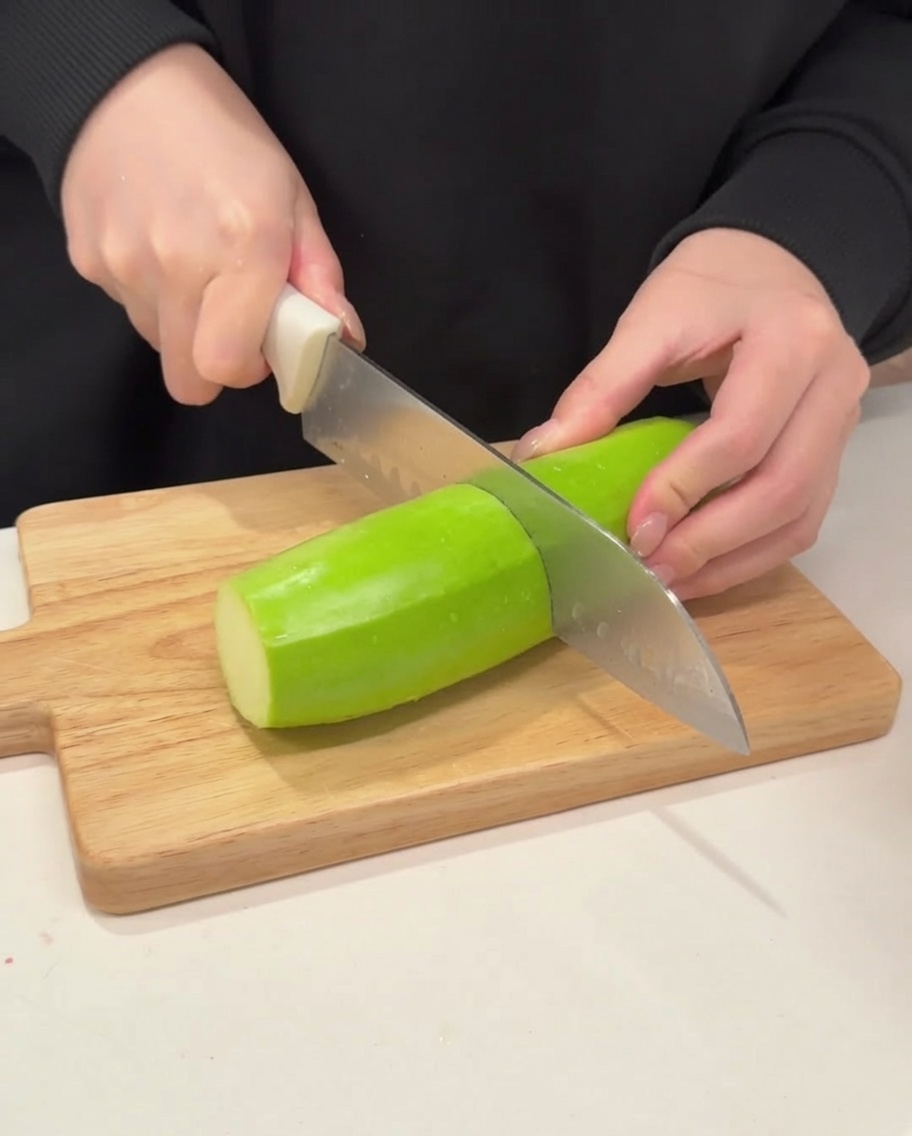 A sharp knife slicing a firm, vibrant green zucchini into thick half-moon shapes on a clean wooden cutting board.