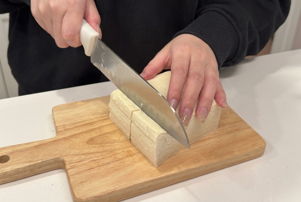 A close-up of a knife slicing a block of firm tofu into cubes on a wooden cutting board.
