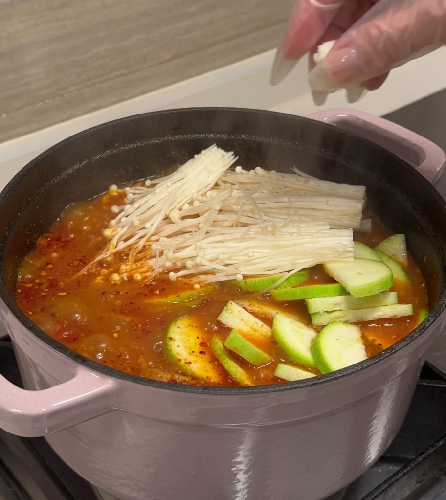 Zucchini slices, tofu cubes, and enoki mushrooms being added to a pot of red, bubbling kimchi broth.
