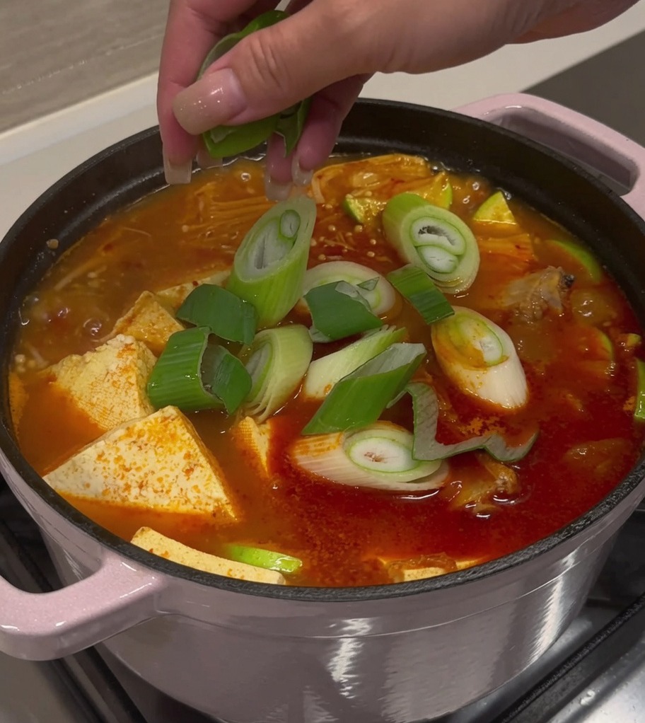 Freshly sliced green onions and chili peppers being sprinkled over a pot of finished kimchi pork rib soup.