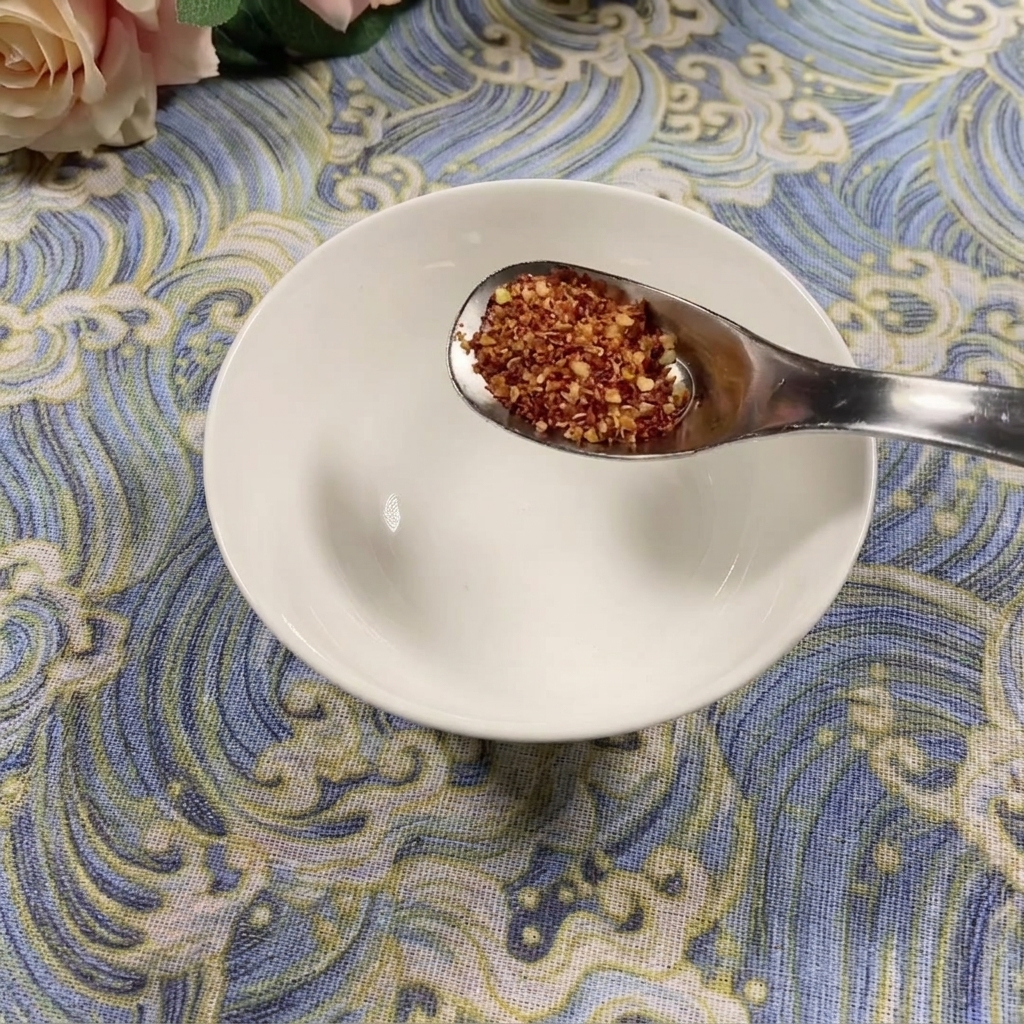 A spoonful of coarse chili powder held over an empty white bowl on a blue patterned surface.