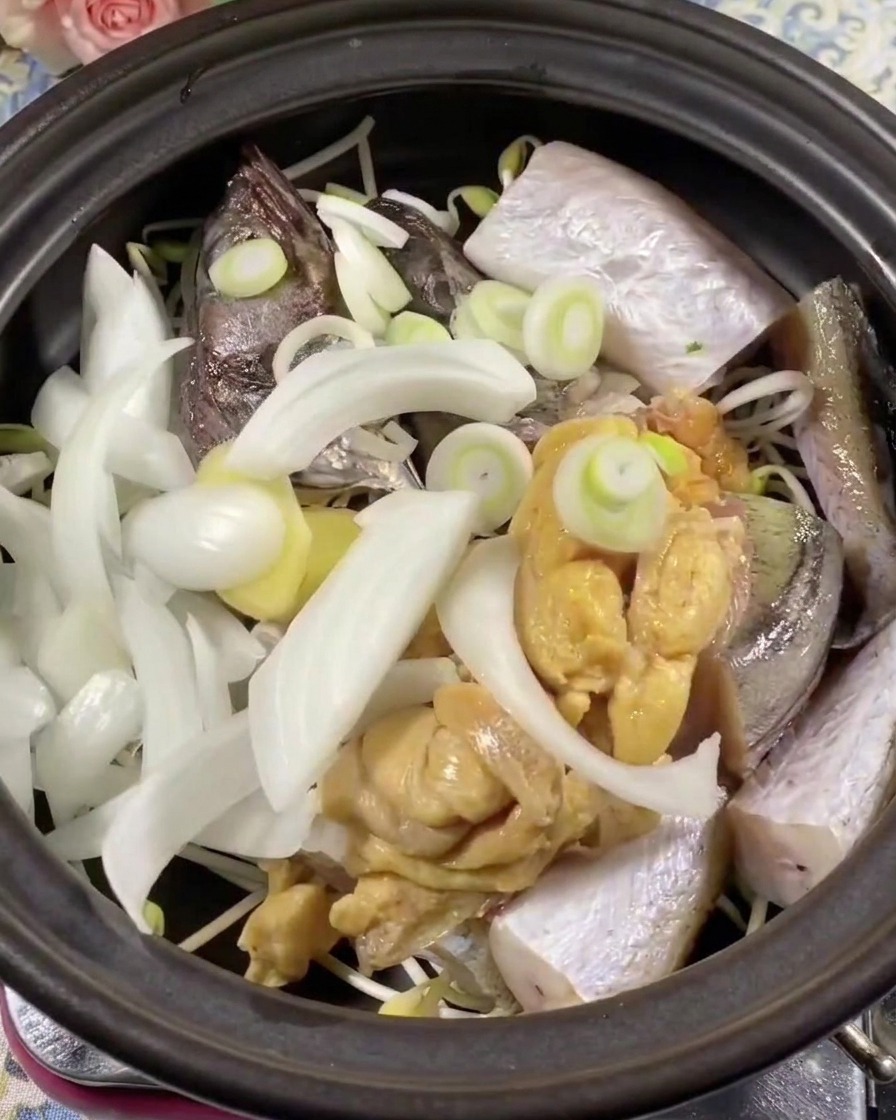 Close-up of green and red chili pepper slices being placed on top of the fish and onions in the clay pot.