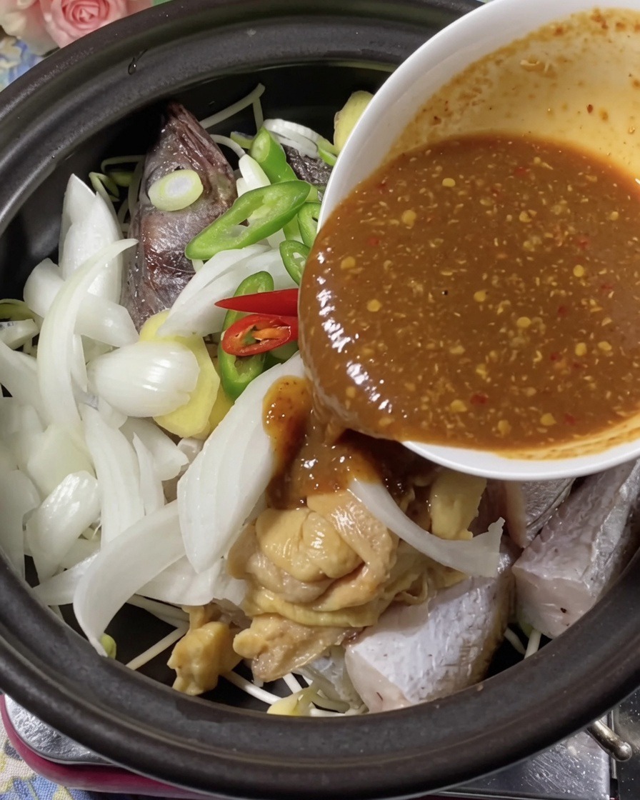 Rich, dark seasoning sauce being poured from a bowl over fish and aromatics in a clay pot.