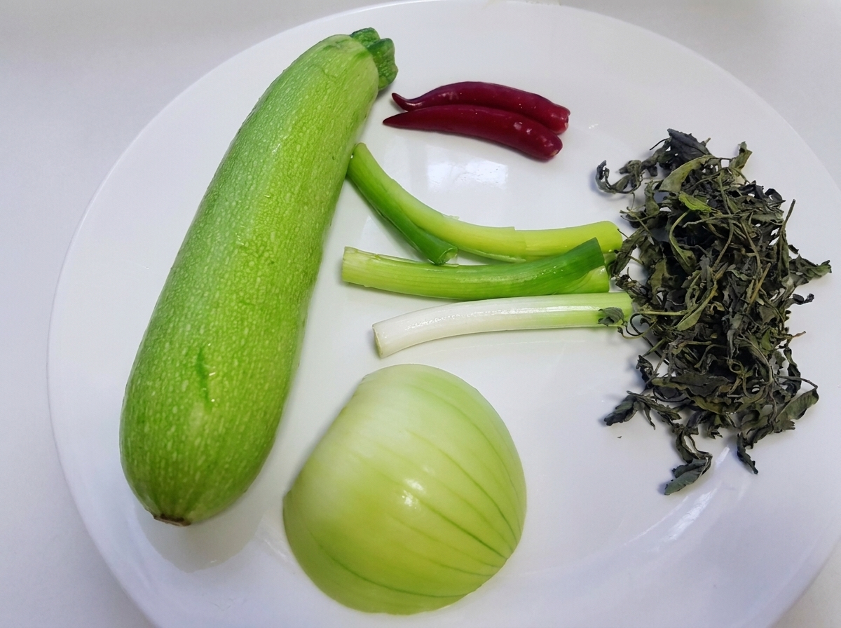 Raw ingredients including a whole green squash, green onions, white onion, wild perilla leaves, and red chili peppers are displayed on a white plate.cite: 1