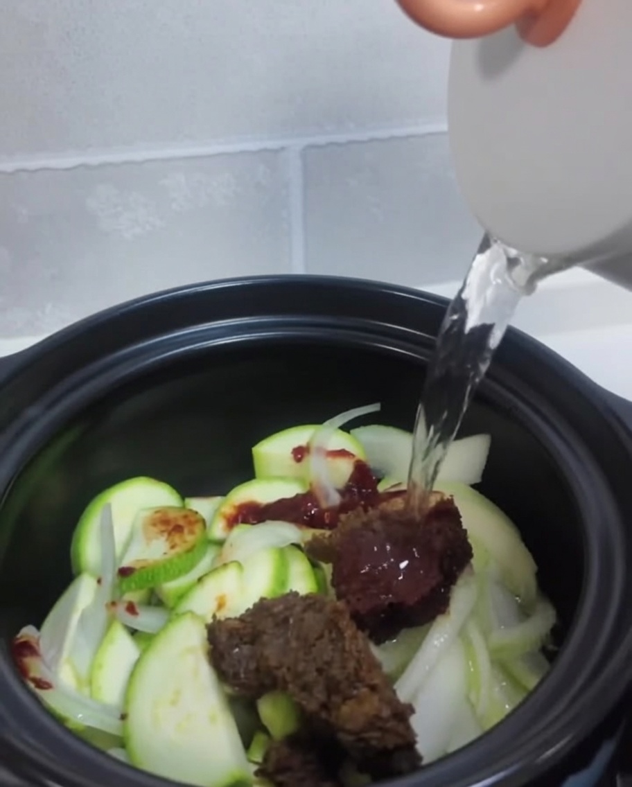 Clear water is poured from a white kettle into the pot over the raw vegetables and seasoning pastes.cite: 1