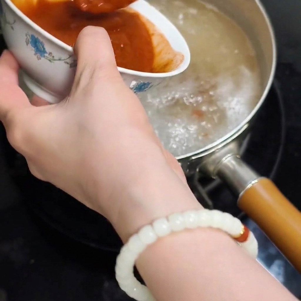 The smooth, red dissolved paste mixture being poured from a bowl into the boiling water in the cooking pot.