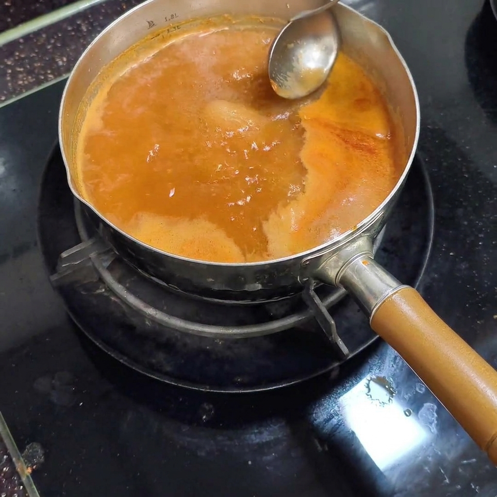 A metal ladle removing light-colored foam from the surface of the simmering red broth.