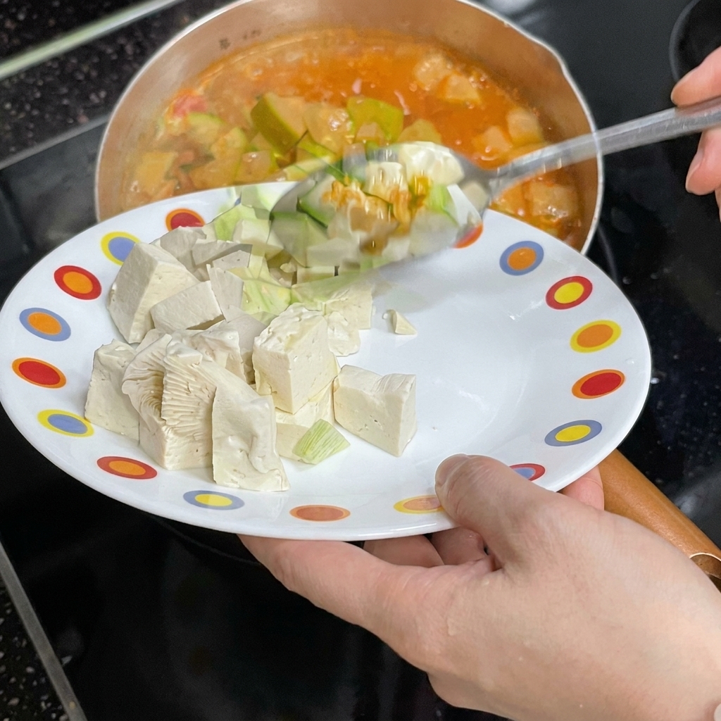 A person sliding white tofu cubes from a colorful plate into a pot of boiling red stew.