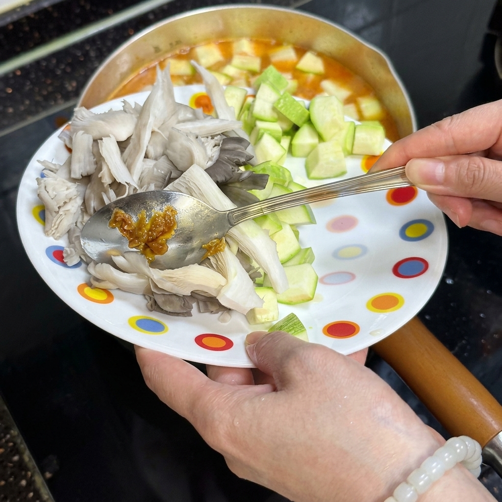 A hand using a spoon to slide diced green zucchini and torn oyster mushrooms from a plate into a bubbling pot.