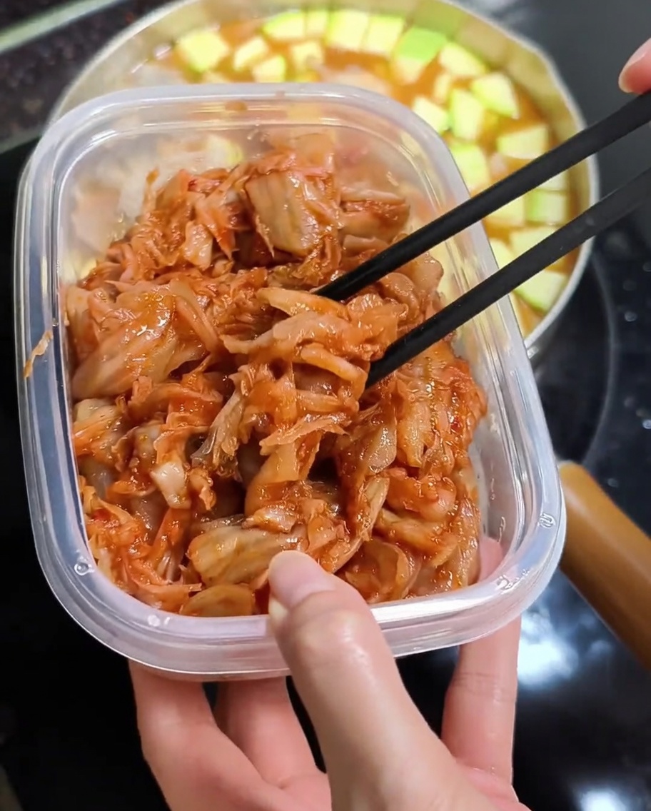 Chopsticks lifting bright red chopped kimchi from a plastic container over a pot of hot stew.